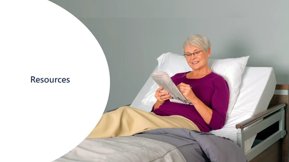 Older woman with short gray hair and glasses sits up in a hospital bed, smiling as she reads a newspaper. A white circle to the left highlights "Resources," offering helpful support during her stay.