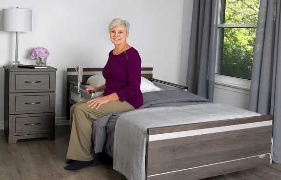 An older woman sits on the edge of a bed in a neatly arranged bedroom with gray furnishings, holding a bed assist rail.