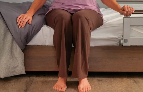 A person sits on the edge of a bed, feet on the floor, holding onto the bedsheet and a bed rail.